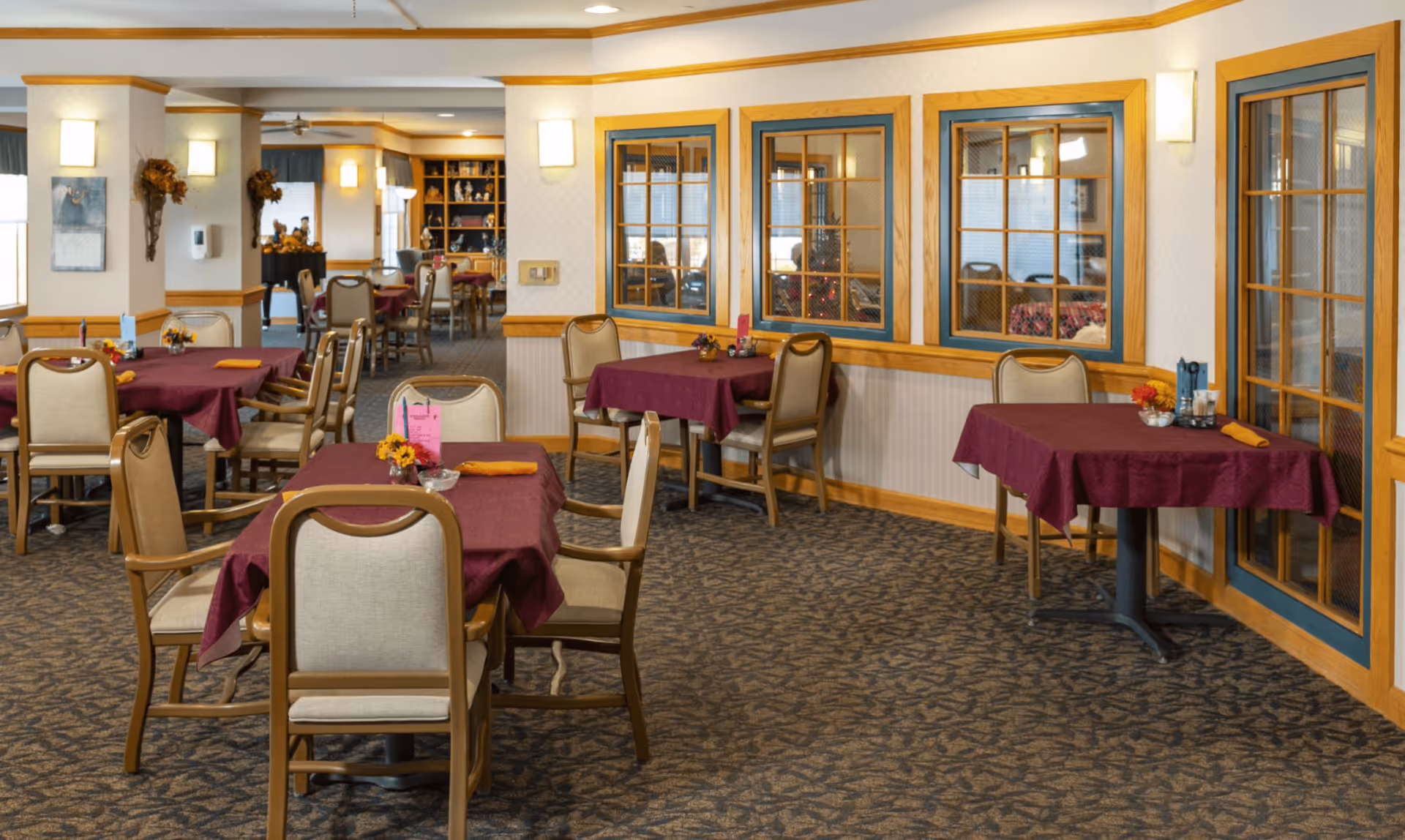 Interior view of a dining room in an assisted living facility with multiple tables covered in burgundy tablecloths and beige cushioned chairs. The room features carpeted flooring, wood trim around windows and walls, and soft wall lighting. Decorative items and a bookshelf are visible in the background.