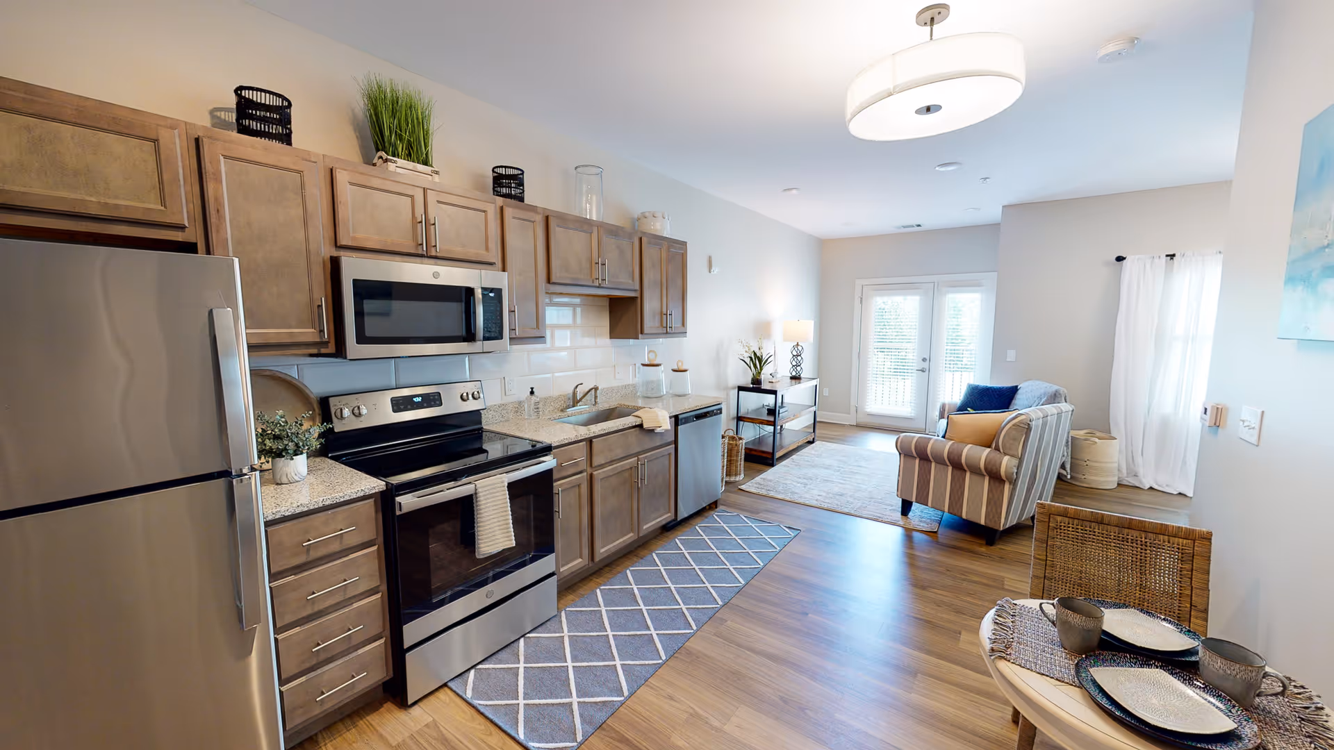 A modern open-concept kitchen and living area featuring stainless steel appliances including a refrigerator, stove, microwave, and dishwasher. The kitchen has wooden cabinets, a granite countertop, and a gray patterned runner rug on the wooden floor. The living area includes a striped armchair, a small table with two chairs set with plates and cups, a console table with a lamp and plant, and large windows with white curtains letting in natural light.