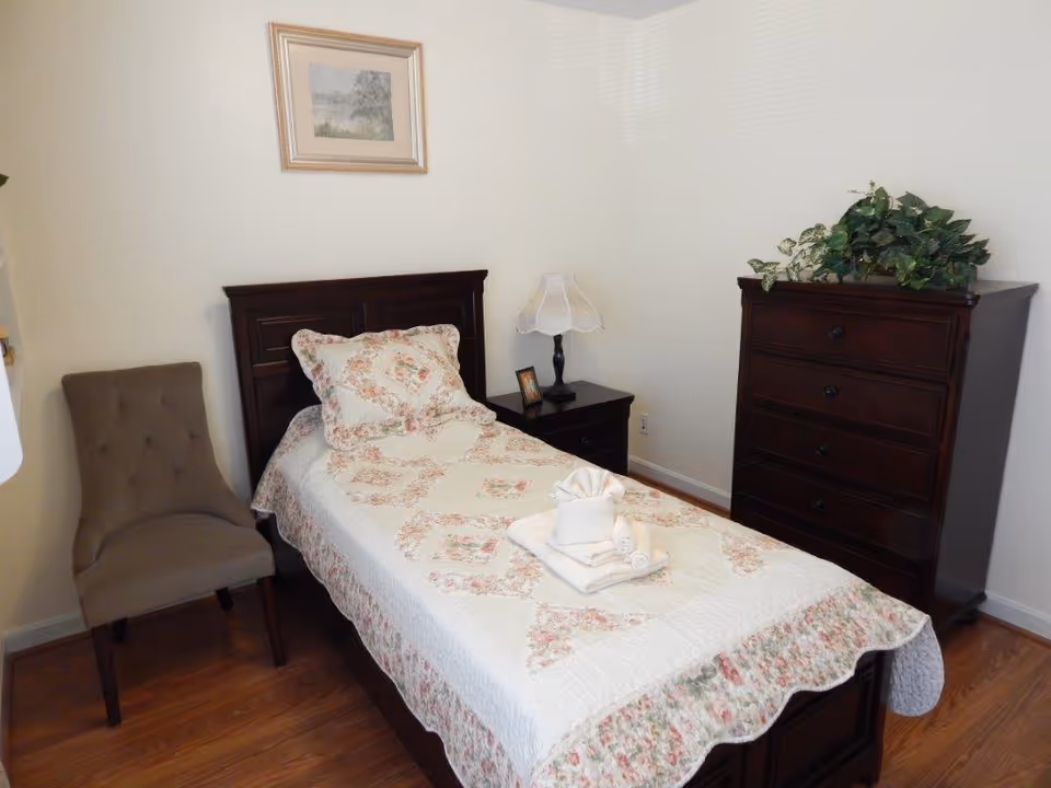 A small bedroom with a single bed covered in a floral quilt and matching pillow. On the bed are neatly folded towels with a towel folded into a decorative shape on top. Next to the bed is a dark wooden nightstand with a lamp and a small framed photo. To the right is a dark wooden chest of drawers with a green leafy plant on top. A beige upholstered chair is positioned to the left of the bed. The walls are light-colored and there is a framed picture hanging above the bed.