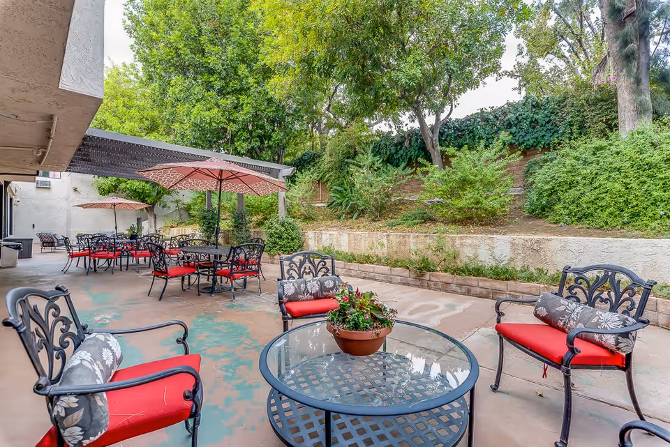 Outdoor patio area with multiple black metal chairs and tables featuring red cushions and floral pillows. Some tables have red umbrellas providing shade. The patio is surrounded by greenery, including trees and bushes, with a retaining wall in the background.