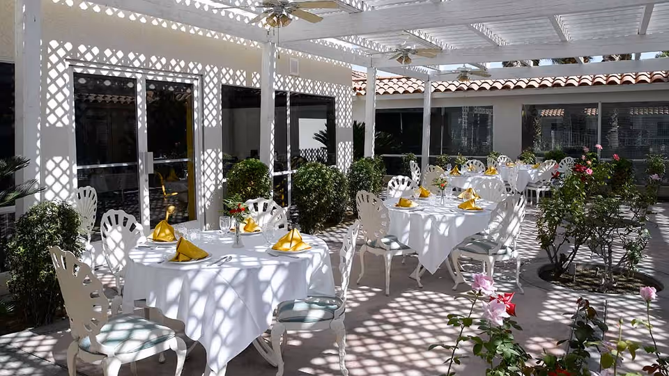Outdoor dining area with several round tables covered in white tablecloths and set with yellow napkins and glassware. White chairs surround the tables, and the area is shaded by a white lattice pergola casting patterned shadows. There are green bushes and blooming flowers around the seating area, with a building featuring large windows and a tiled roof in the background.
