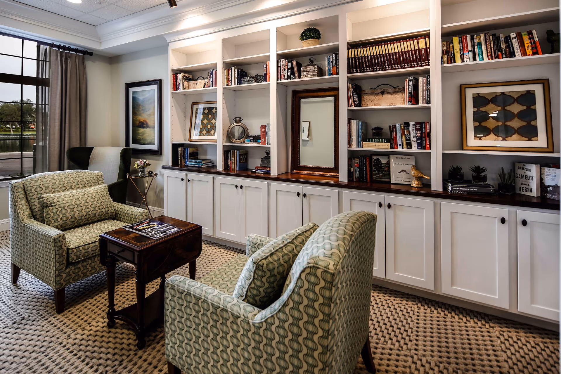A cozy sitting area with two patterned armchairs and a small wooden table between them, positioned in front of a built-in white bookshelf filled with books, framed pictures, and decorative items. A large window with curtains allows natural light to enter the room, and a framed painting hangs on the wall beside the window.