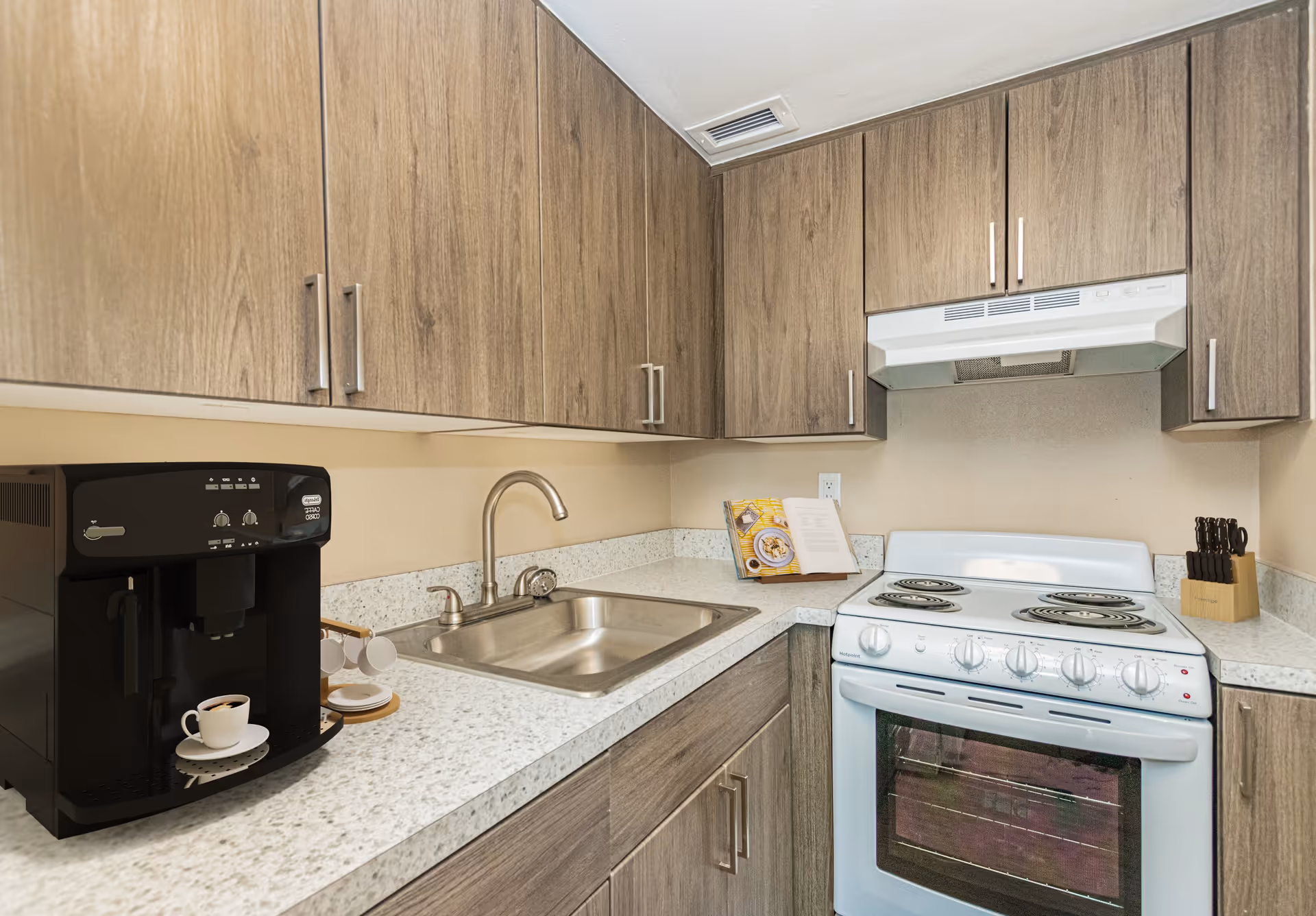 Compact kitchen with wood cabinets, a countertop coffee machine beside a stainless sink, and a white electric stove with range hood.