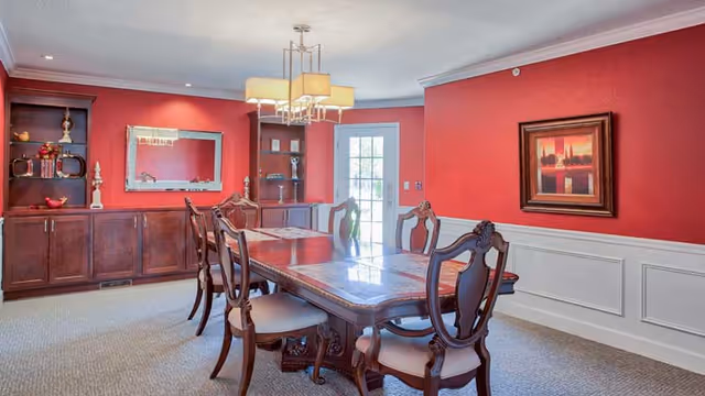 A dining room with a long wooden dining table and six matching chairs with cushioned seats. The walls are painted red with white wainscoting, and there is a built-in wooden cabinet with shelves and decorative items on the left side. A rectangular mirror hangs above the cabinet. A modern chandelier with rectangular shades hangs above the table. A framed painting is on the right wall, and a glass door leading outside is visible in the background.