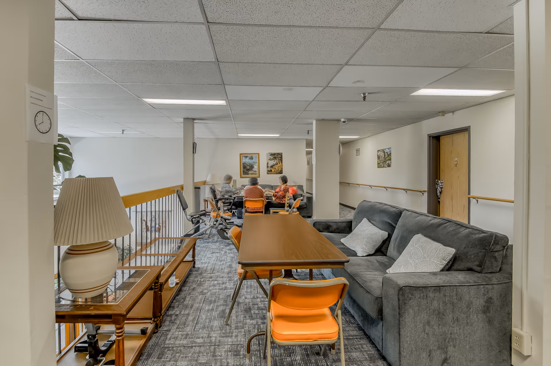 A common area in a senior living facility with a gray couch, a wooden table with orange chairs, a lamp on a side table, and three elderly people sitting and conversing in the background near framed pictures on the wall.