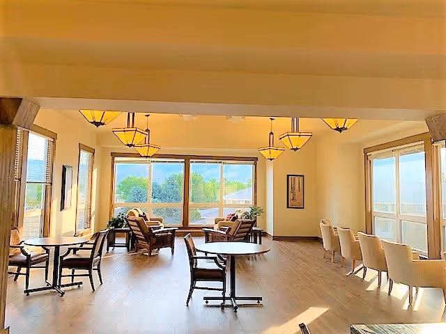 A bright and spacious common area in an assisted living facility with large windows letting in natural light. The room features multiple round tables with chairs, a seating area with armchairs near the windows, wooden flooring, and several hanging light fixtures. The walls are light-colored, and there are framed pictures and plants adding to the decor.