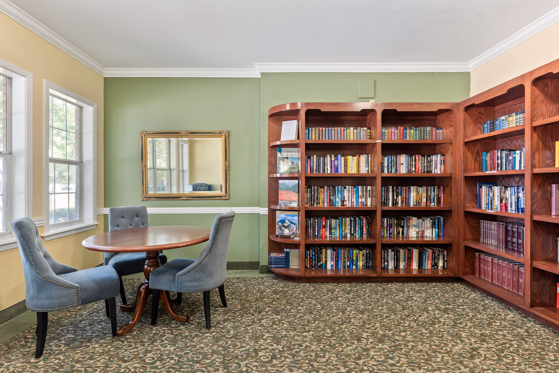 A cozy reading room with a round wooden table surrounded by three blue upholstered chairs. The room features large windows letting in natural light, a green and yellow painted wall with a decorative mirror, and a large wooden bookshelf filled with books.