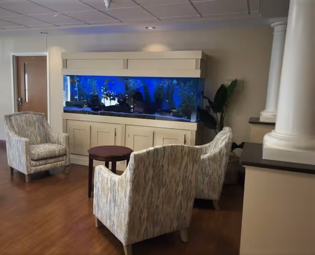 A cozy seating area in a senior living facility with three patterned armchairs arranged around a small round wooden table. Behind the chairs is a large built-in aquarium filled with water, plants, and fish. The room has wooden flooring, beige walls, and decorative white columns on the right side.