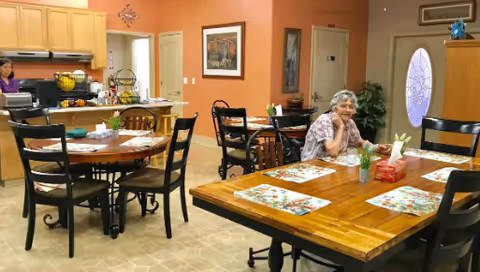 A cozy dining area in an assisted living facility with wooden tables and black chairs. An elderly woman is sitting at one of the tables, smiling. The room has a kitchen area in the background with light wood cabinets and a countertop. The walls are painted a warm orange color and decorated with framed pictures and a clock. There is a door with a decorative glass window and some plants near it.