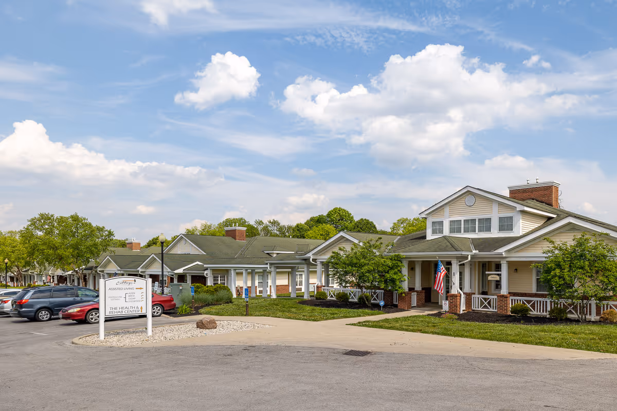 Exterior view of a single-story assisted living and health rehab center building with a covered entrance, American flag, and a parking lot with several cars. The sky is partly cloudy with blue patches.