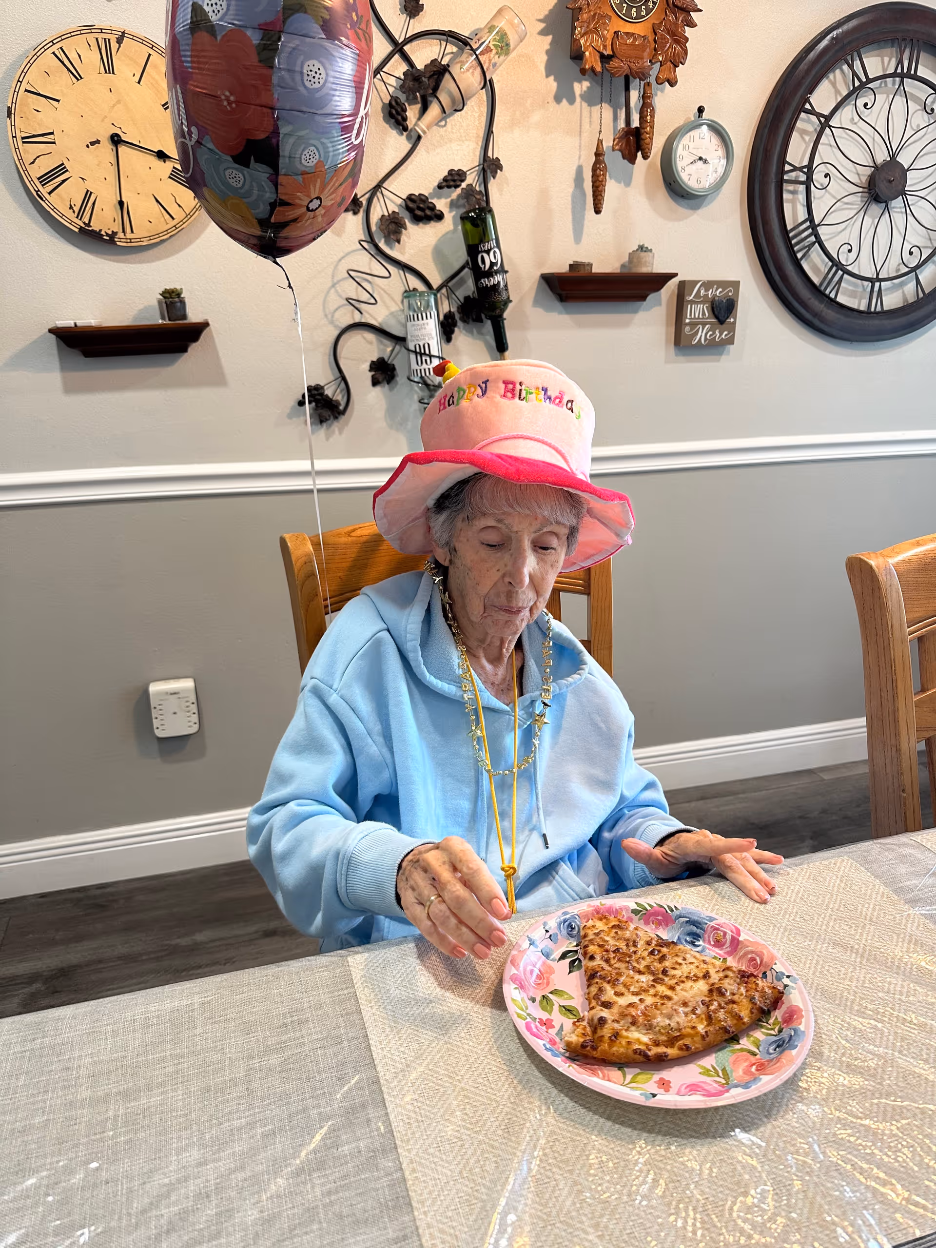 An elderly woman wearing a "Happy Birthday" hat sits at a dining table with a slice of pizza on a floral plate and a balloon nearby in a decorated room.