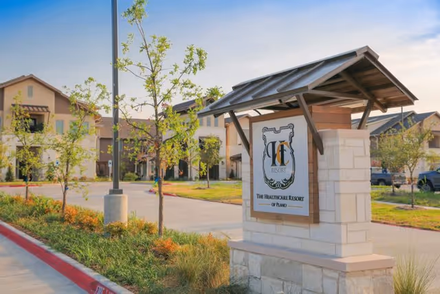 Outdoor view of the entrance sign for The Healthcare Resort of Plano, featuring a stone and wood structure with the facility's logo and name. The background shows a paved driveway, landscaped greenery, young trees, and residential-style buildings under a clear sky.