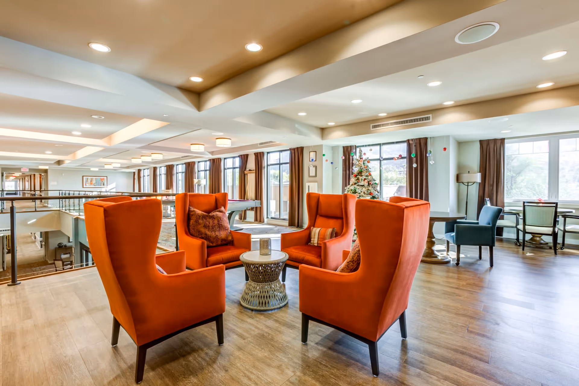 A bright and spacious common area with four orange high-back armchairs arranged around a small round table. The room features large windows with brown curtains, wooden flooring, and a decorated Christmas tree near the windows. Additional seating and tables are visible in the background, along with ceiling lights and a railing overlooking a lower level.