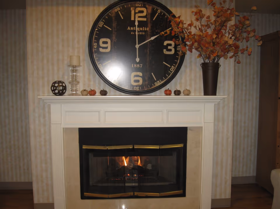 A cozy indoor fireplace with a white mantel decorated with small pumpkins, a candle holder, a decorative sphere, and a tall vase with autumn leaves. Above the mantel hangs a large black clock with gold numbers and hands.