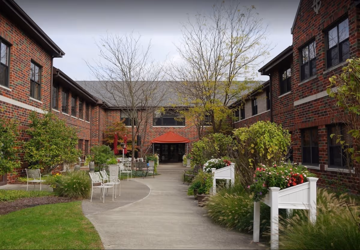 Outdoor courtyard area at Concordia at Villa St. Joseph with a paved walkway, benches, planters with flowers, and brick buildings on both sides under a cloudy sky.