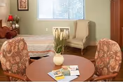 Cozy senior living lounge with round table, upholstered chairs, brochures, vase, and bed near a window.