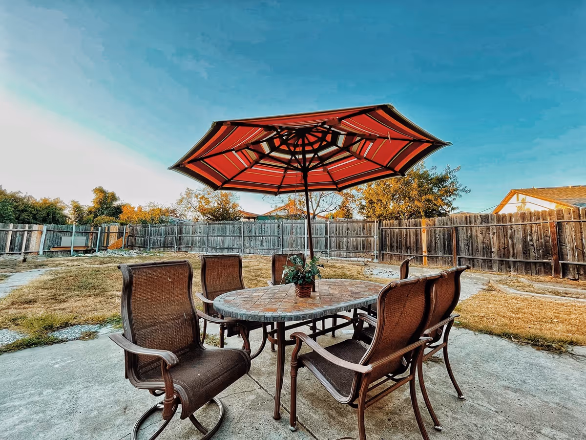 Outdoor patio area with a round table and six brown chairs under a large red and white striped umbrella. The patio is surrounded by a wooden fence and there is grass and trees in the background under a clear blue sky.