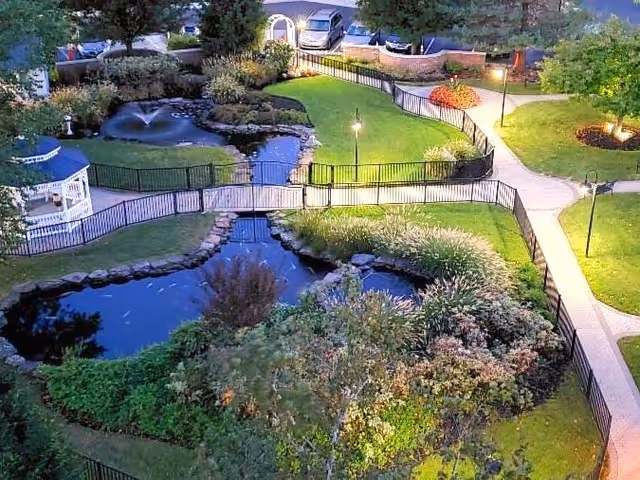 A well-maintained outdoor garden area at dusk featuring a small pond with a fountain, a white gazebo, stone-bordered water features, green lawns, paved walking paths, black metal fencing, and several trees and shrubs. There are also parked cars visible in the background.