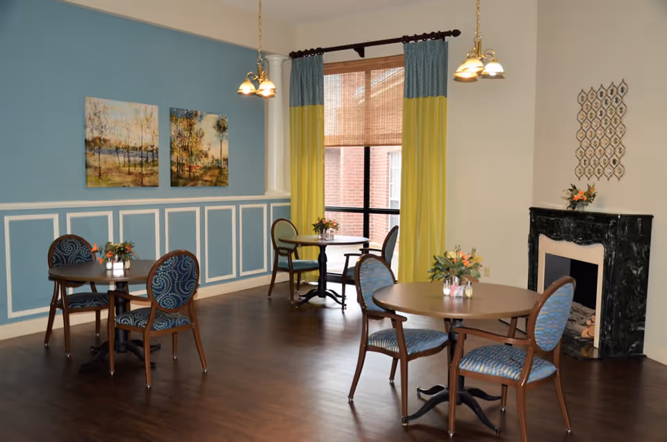 A cozy dining area with three round wooden tables, each surrounded by two upholstered chairs with blue patterned fabric. The room features a blue accent wall with white trim, two landscape paintings, a window with yellow and blue curtains, and a black marble fireplace decorated with a small flower arrangement. The floor is dark wood, and two hanging light fixtures illuminate the space.