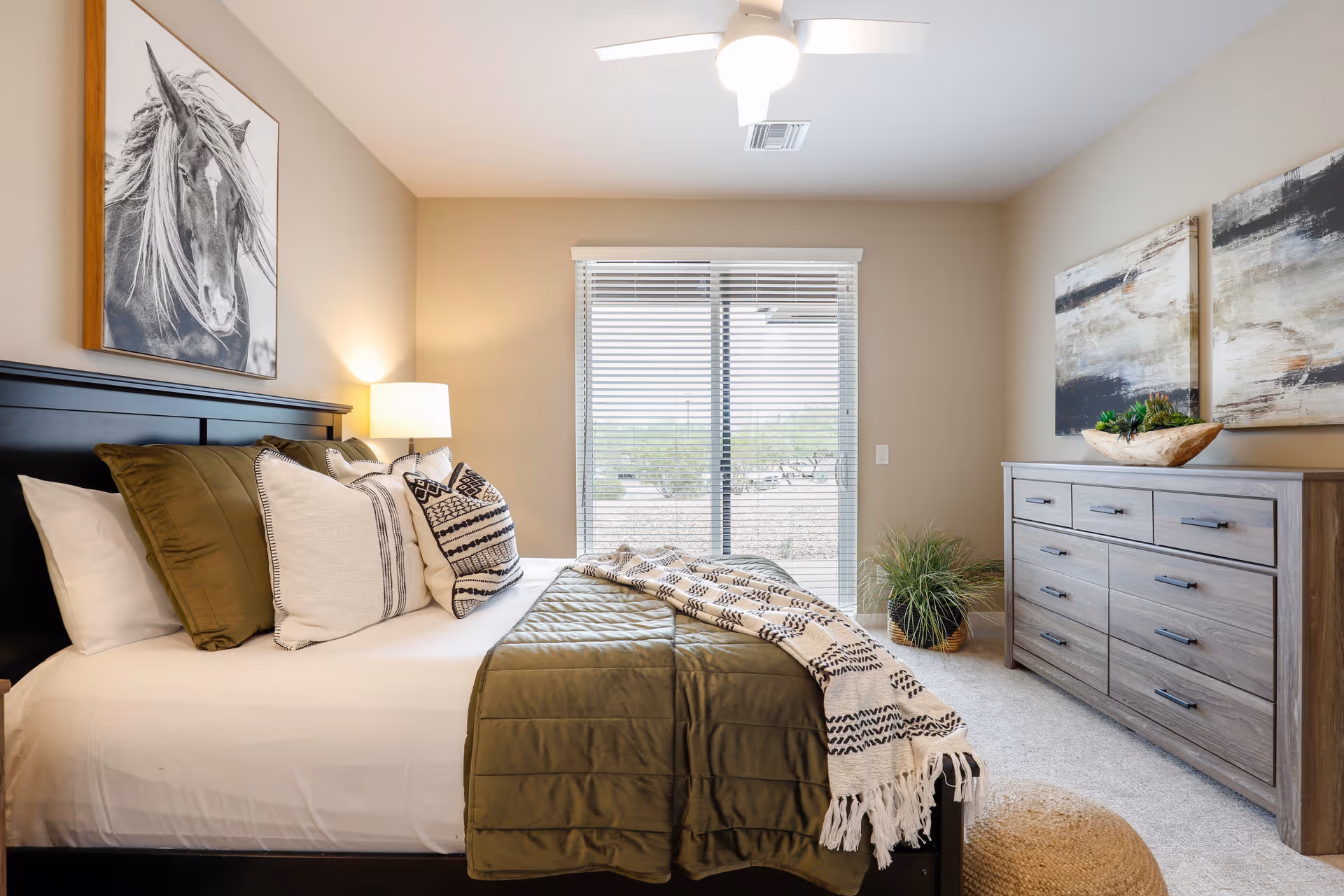 A neatly made bed with green and white pillows and a green quilt in a bright bedroom. There is a large window with blinds letting in natural light, a wooden dresser with decorative plants and abstract paintings above it, a ceiling fan with light, and a framed black and white horse picture on the wall.