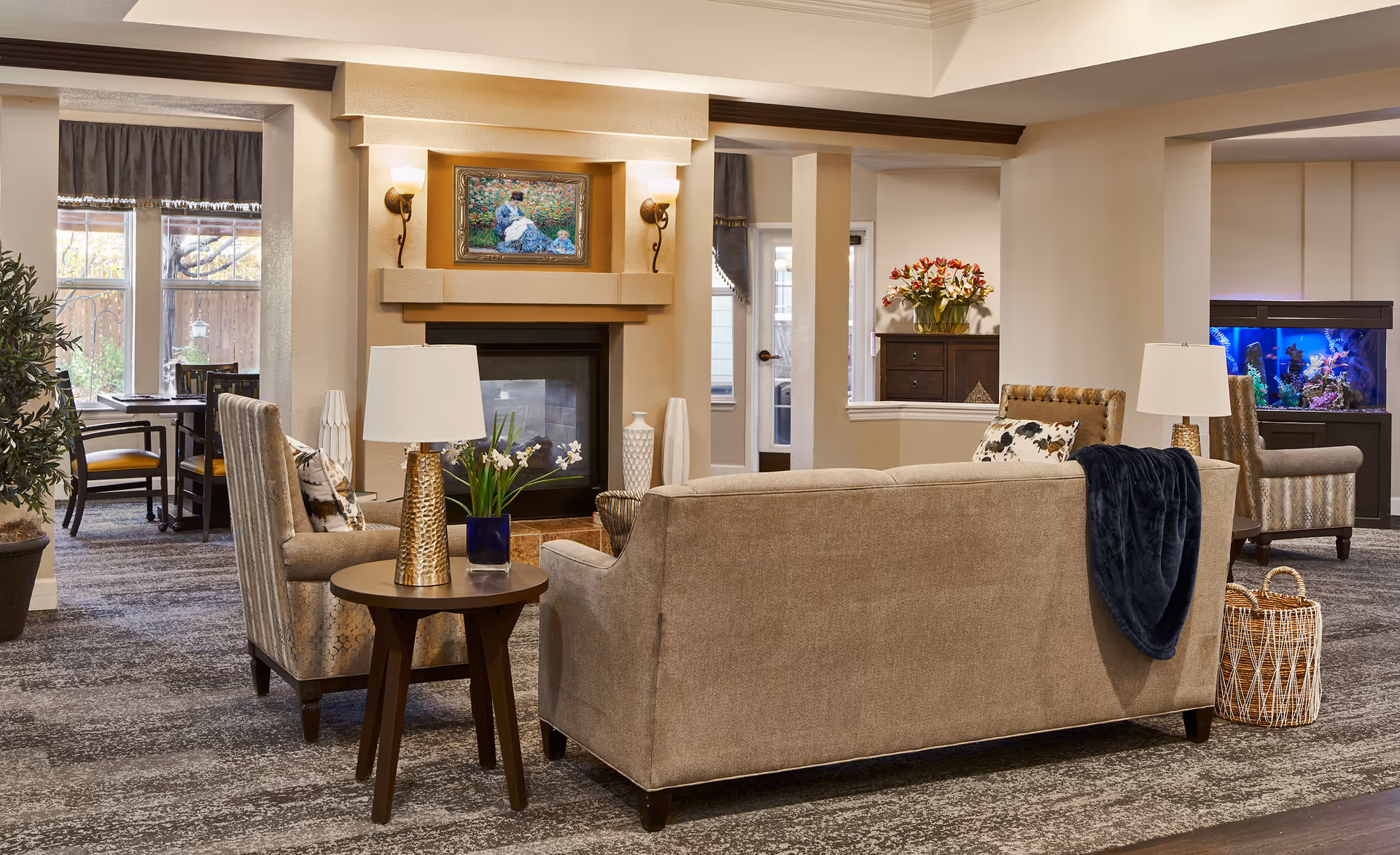A cozy living room area in an assisted living facility featuring beige sofas and armchairs arranged around a fireplace with a painting above it. There are side tables with lamps and decorative plants, a large window with curtains, and a fish tank in the background. The room has a warm and inviting atmosphere with neutral tones and soft lighting.