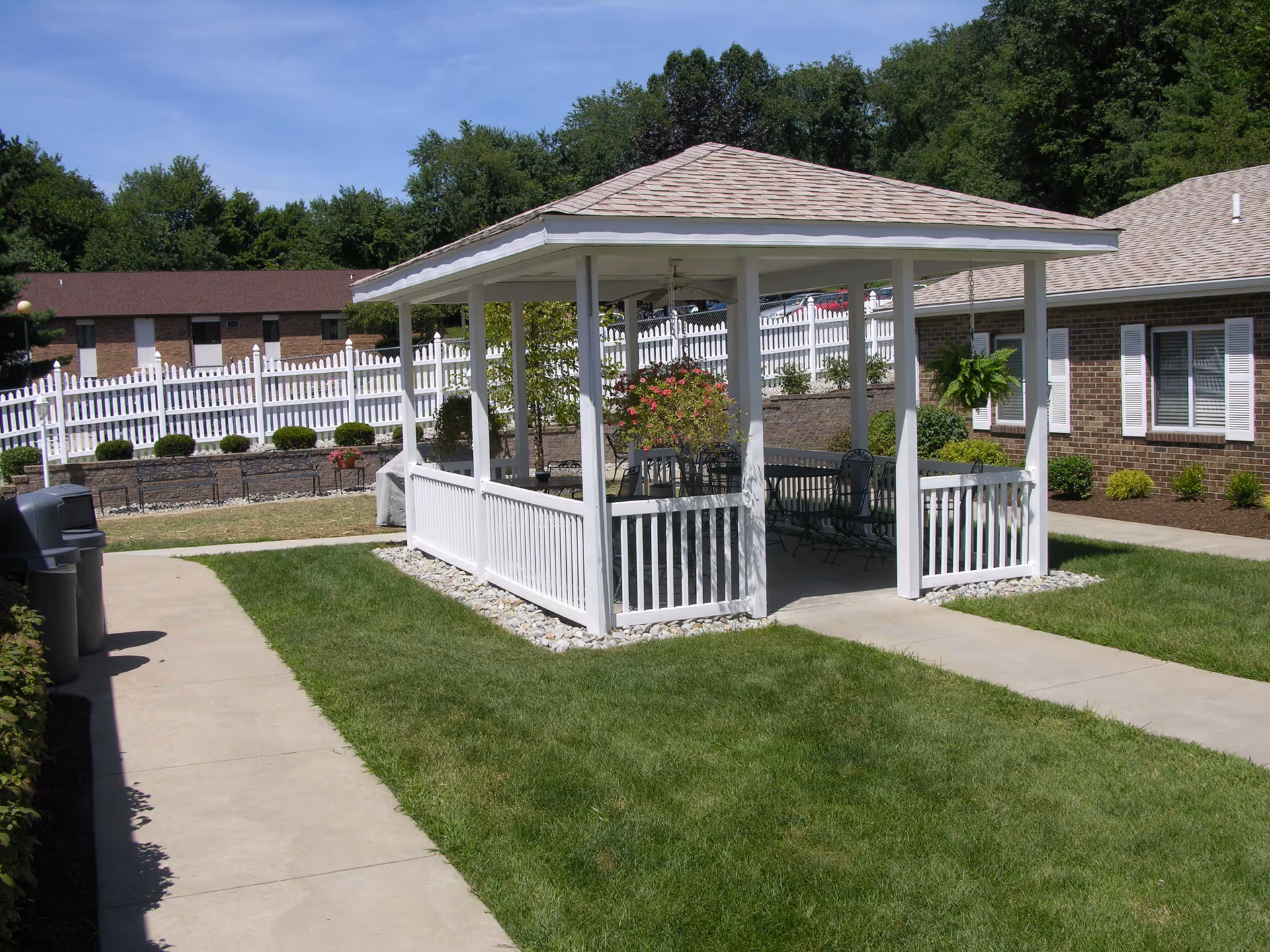 White wooden gazebo with seating in a grassy courtyard surrounded by a white picket fence and brick buildings.