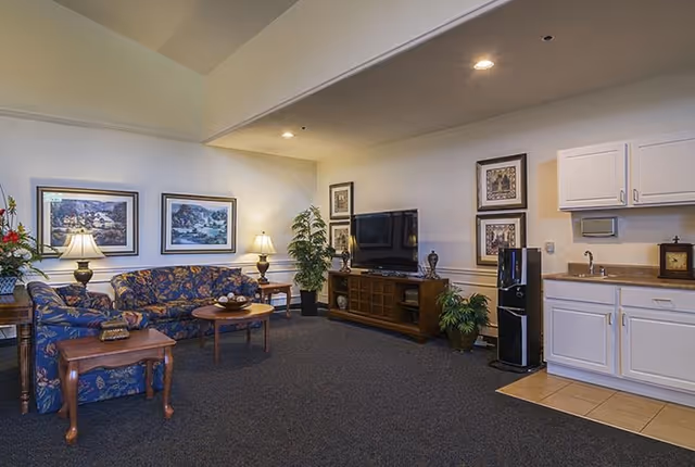 A cozy living room area in a senior living facility featuring a floral patterned sofa and armchair, two wooden side tables with lamps, framed landscape artwork on the walls, a large flat-screen TV on a wooden TV stand, potted plants, and a small kitchenette with white cabinets and a water dispenser.