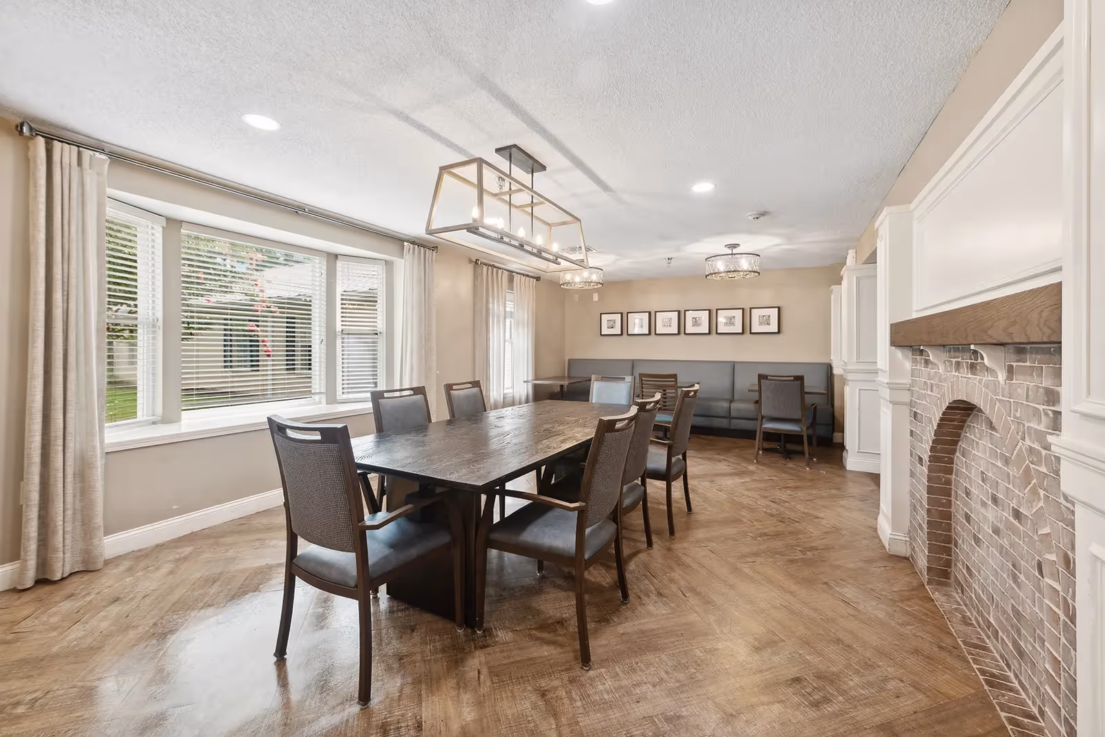 Communal dining room with a long wooden table surrounded by chairs, banquette seating along the back wall, a large window, and overhead lighting.