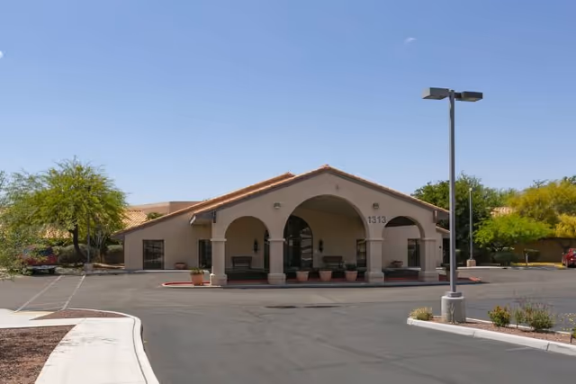 One-story stucco building with arched covered entrance, parking lot, and desert landscaping under a clear sky.