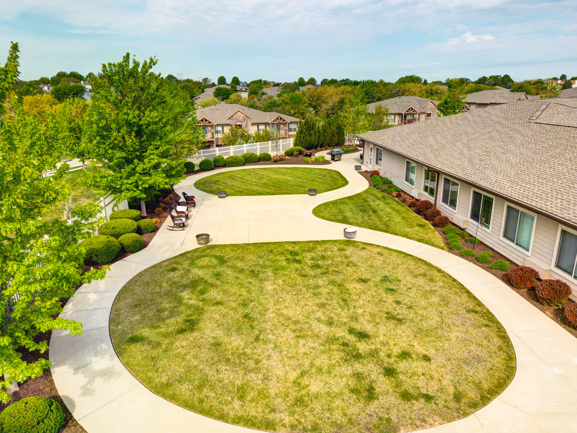A landscaped outdoor area at Cedar Creek of Prairie Meadows featuring a large circular grassy lawn surrounded by a curved concrete walkway. There are several bushes and trees along the walkway, with a few chairs placed near the trees. The side of a building with multiple windows is visible on the right side, and residential buildings can be seen in the background under a partly cloudy sky.