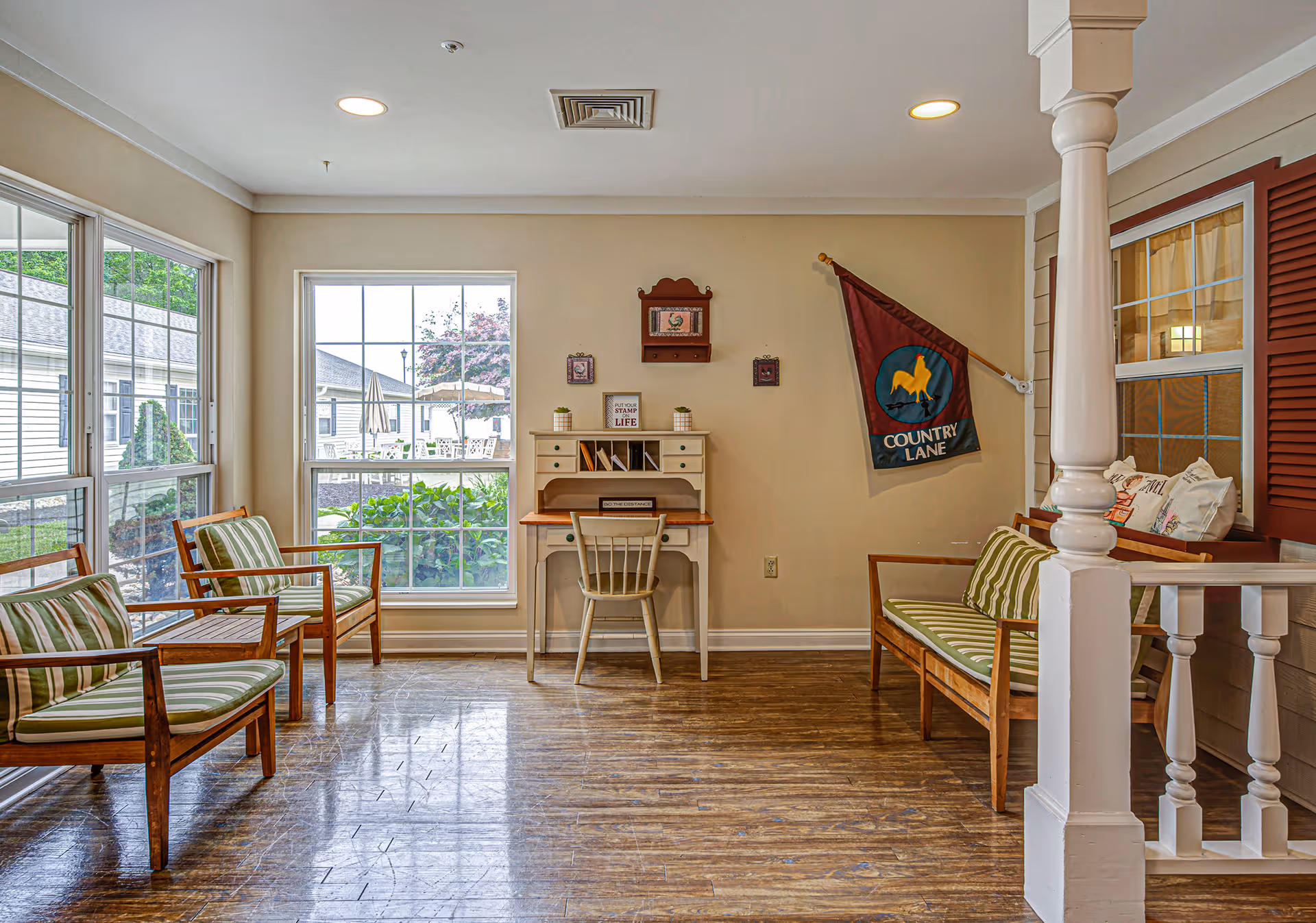 A cozy sitting area with wooden chairs and a bench featuring green and white striped cushions. There is a small white desk with a chair against the wall, decorated with small plants and framed pictures. A flag with a yellow horse and the words 'Country Lane' hangs on the wall. Large windows provide a view of an outdoor patio area with tables and umbrellas.