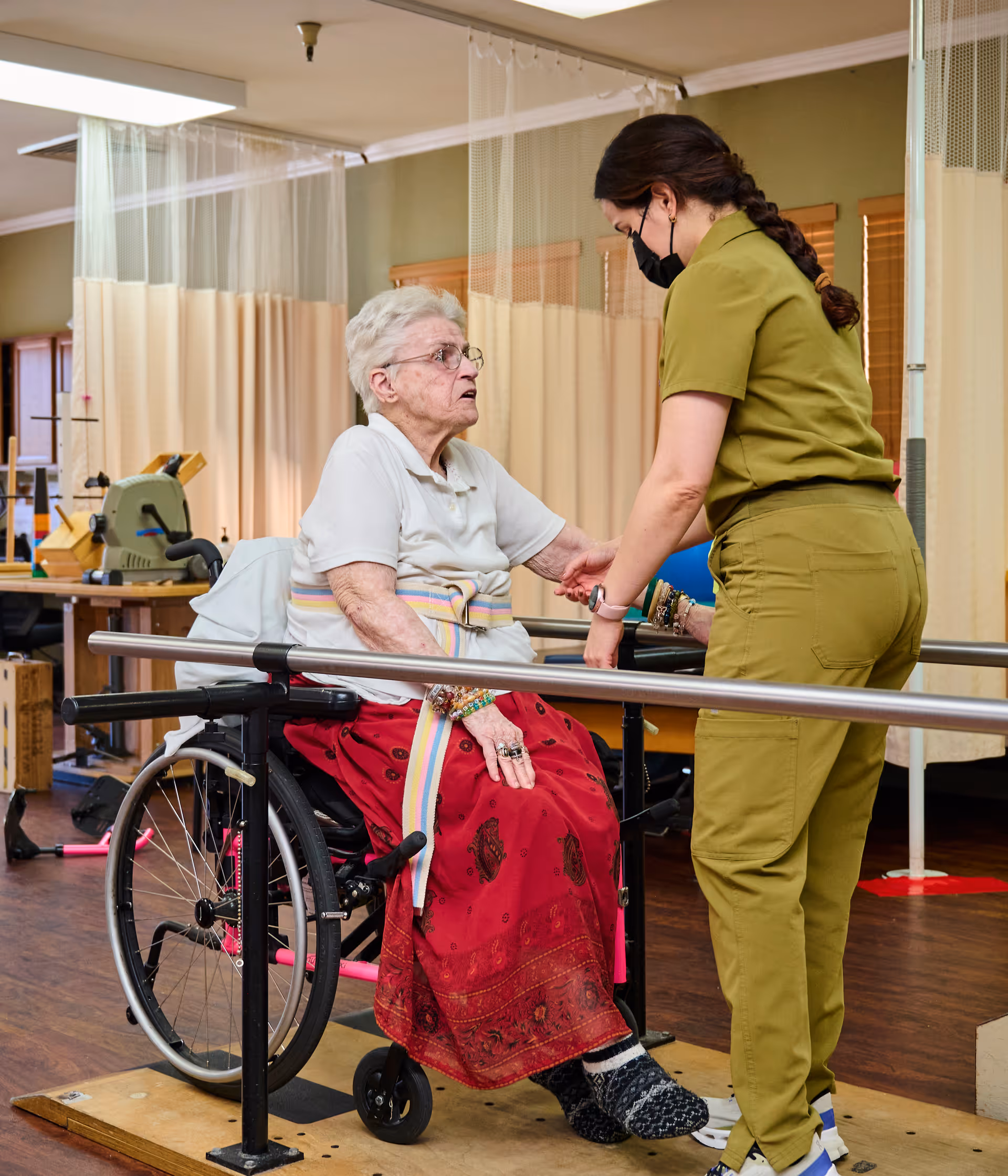 An elderly woman in a wheelchair is assisted by a caregiver wearing a mask and green scrubs in a rehabilitation center. The elderly woman is holding onto parallel bars, and the caregiver is supporting her arm. The room has wooden floors and beige privacy curtains in the background.