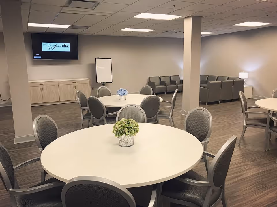 A spacious common area with round tables and chairs arranged for group seating. Each table has a small decorative plant in the center. In the background, there are several gray armchairs arranged along the walls, a flat screen TV mounted on the wall displaying the Birchwood Suites logo, a whiteboard, and a wooden cabinet. The room has wood flooring and neutral-colored walls with ceiling lights providing bright illumination.