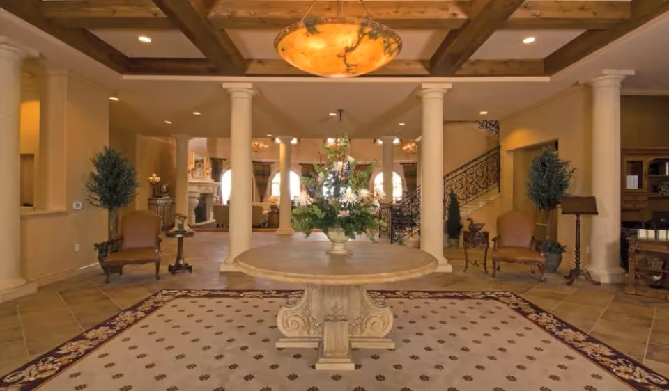 Elegant entry lobby with a round center table topped by a floral arrangement, flanked by columns, chairs, and a staircase.