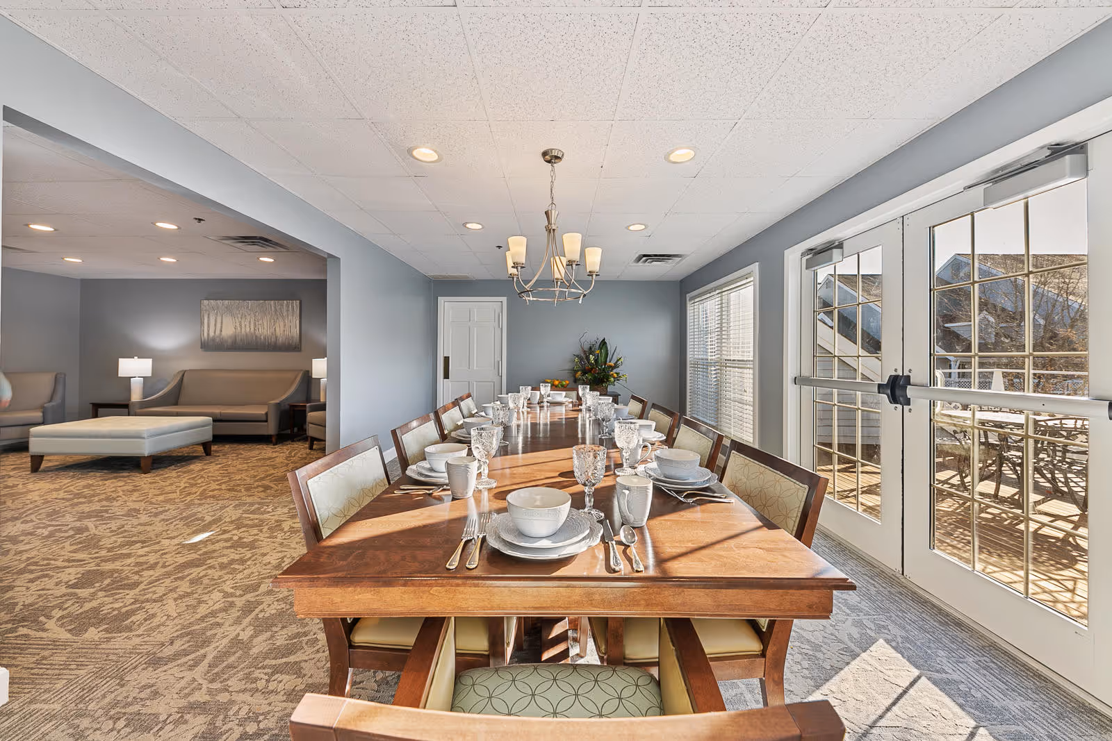 A well-lit dining room with a long wooden table set for a meal with plates, bowls, glasses, and silverware. The room has large windows and glass doors allowing natural light to enter. Adjacent to the dining area is a lounge space with sofas, a large ottoman, and lamps. The walls are painted light gray and the carpet has a patterned design.