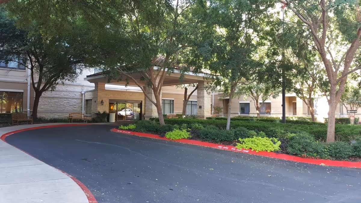 Entrance of a senior living facility with a covered drop-off area, surrounded by trees, bushes, and benches. The building exterior is beige with stone accents and large windows.