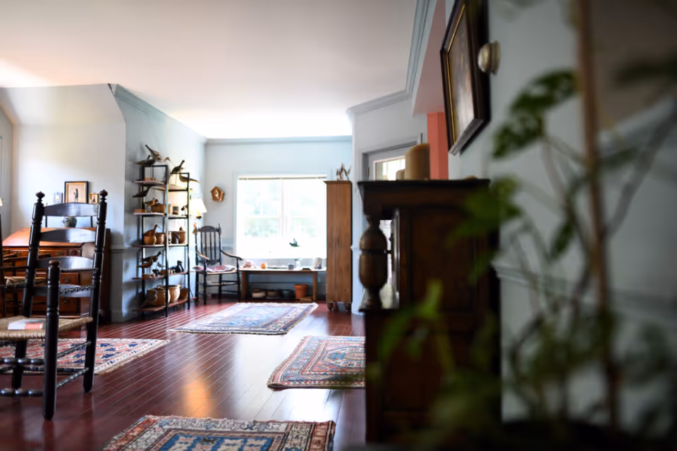 A cozy living room with wooden floors and multiple patterned rugs. There are wooden chairs, a wooden desk, and a shelving unit with various decorative items. A large window lets in natural light, and there are plants and framed pictures on the walls.