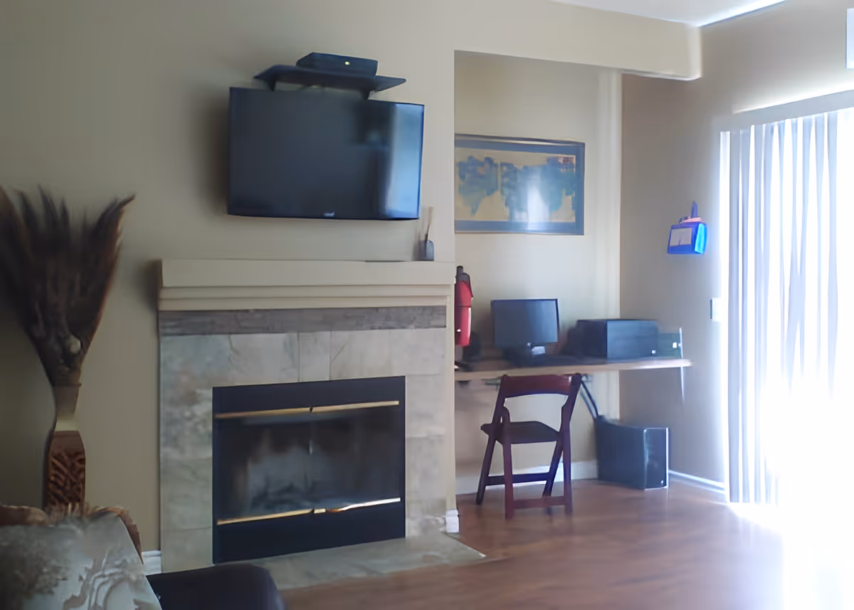 A cozy interior room featuring a wall-mounted flat screen TV above a stone fireplace. To the right, there is a small wooden desk with a chair, a computer monitor, a printer, and a red thermos. A framed artwork hangs on the wall above the desk. Vertical blinds cover a large window or sliding door, allowing natural light to brighten the room. A decorative vase with tall dried plants is placed on the left side near a couch.