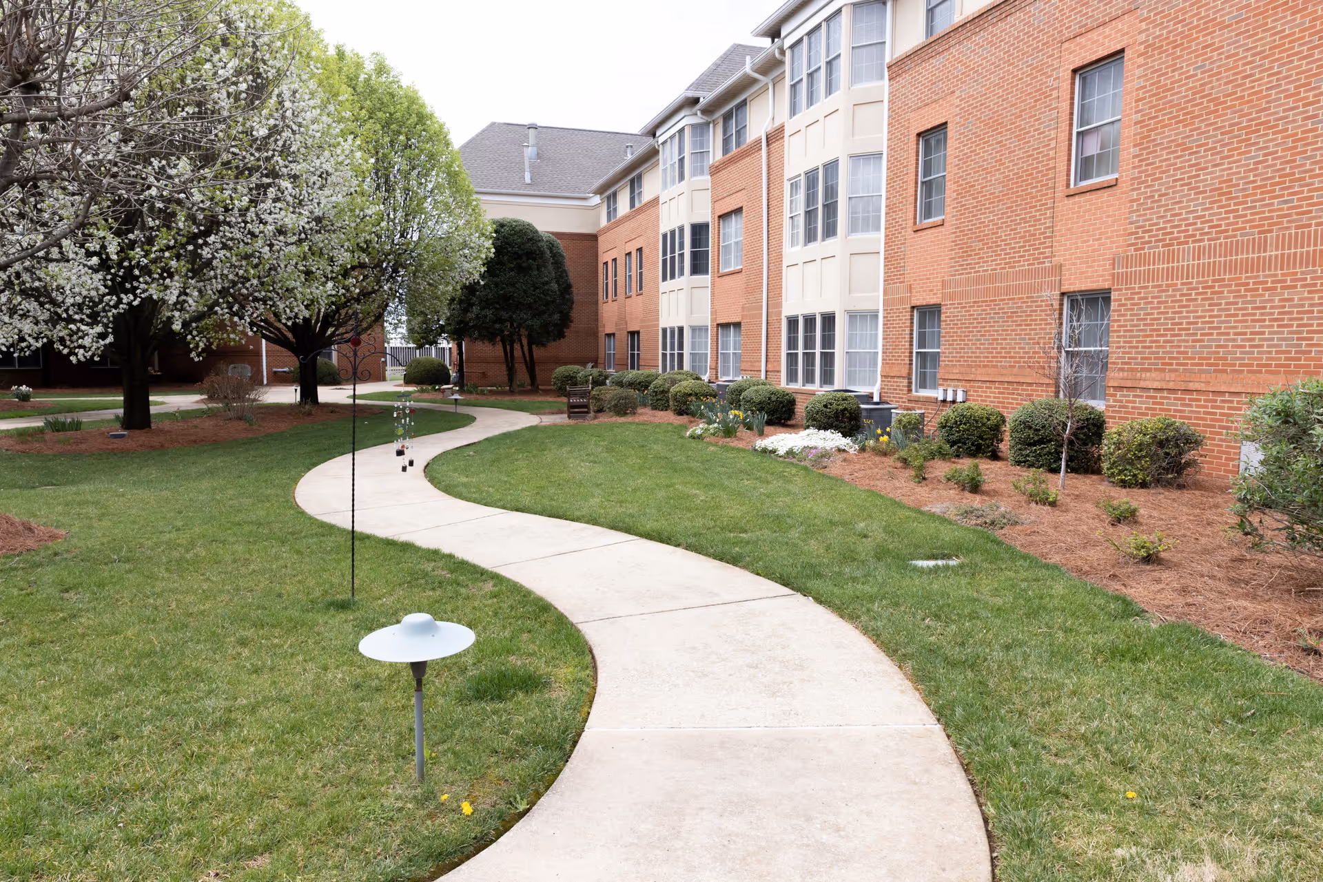 Curved concrete pathway through a well-maintained garden area with green grass, blooming trees, and shrubs alongside a multi-story brick building with many windows.
