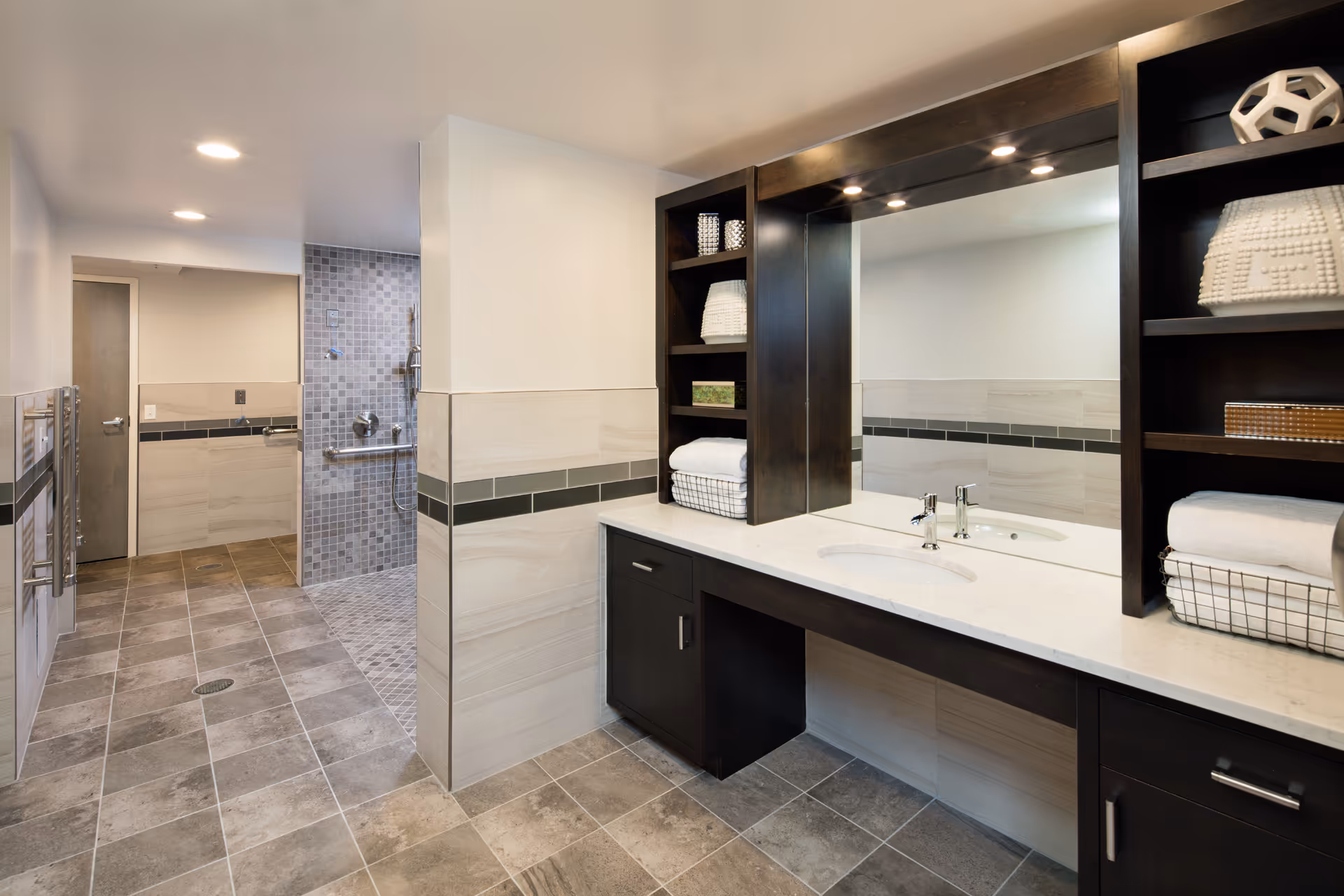 A modern bathroom with a large vanity featuring a white countertop, built-in sink, and dark wood cabinetry with open shelves holding towels and decorative items. The room has tiled floors and walls, recessed lighting, and a walk-in shower area with a tiled wall and grab bars.
