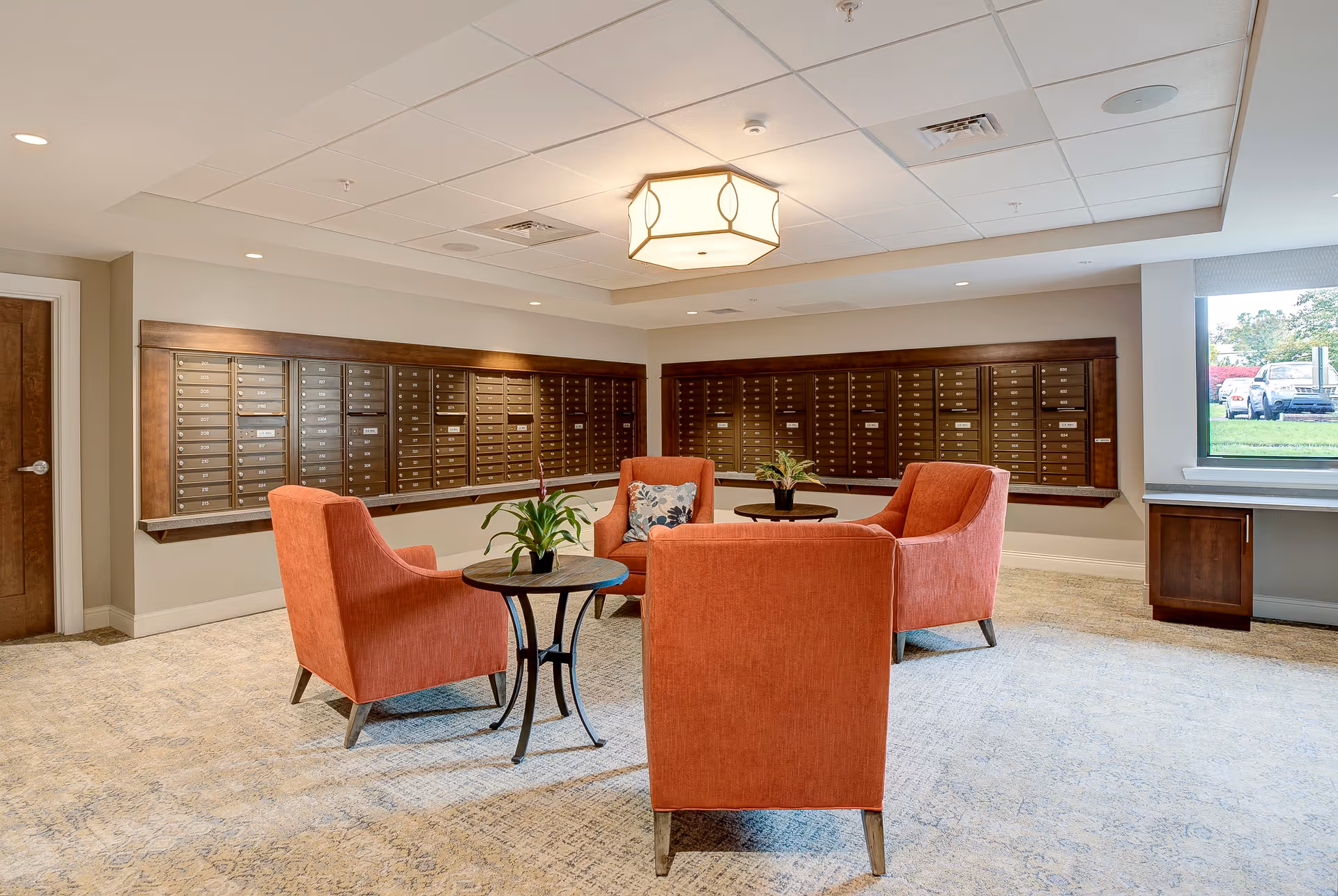 A seating area with four orange armchairs arranged around two small round tables with potted plants. Behind the seating area is a wall with multiple mailboxes. The room has a carpeted floor, a ceiling light fixture, and a window showing a view of parked cars outside.