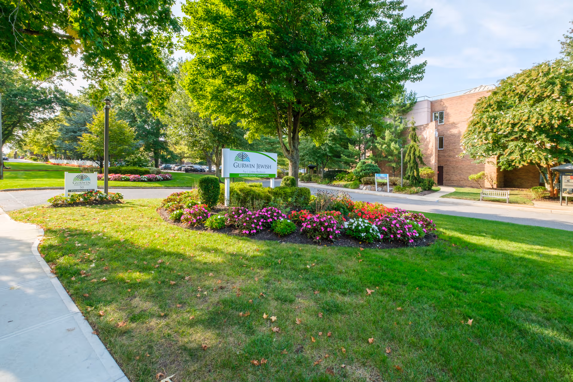 Landscaped front lawn with flower beds, signage reading 'Gurwin Jewish', and the residence building entrance in the background.