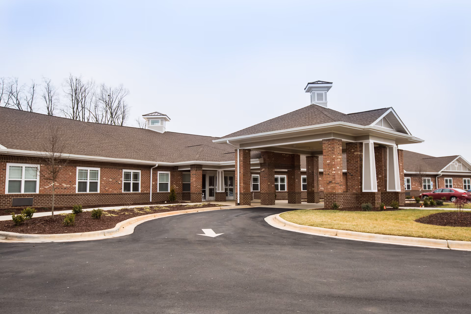 Exterior view of Mebane Ridge Assisted Living facility showing a single-story brick building with a covered entrance driveway, landscaped areas with small shrubs and grass, and a few parked cars in the background under a clear sky.