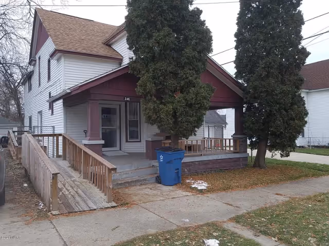 Two-story house with a covered front porch, wooden wheelchair ramp, and two large evergreen trees in front.