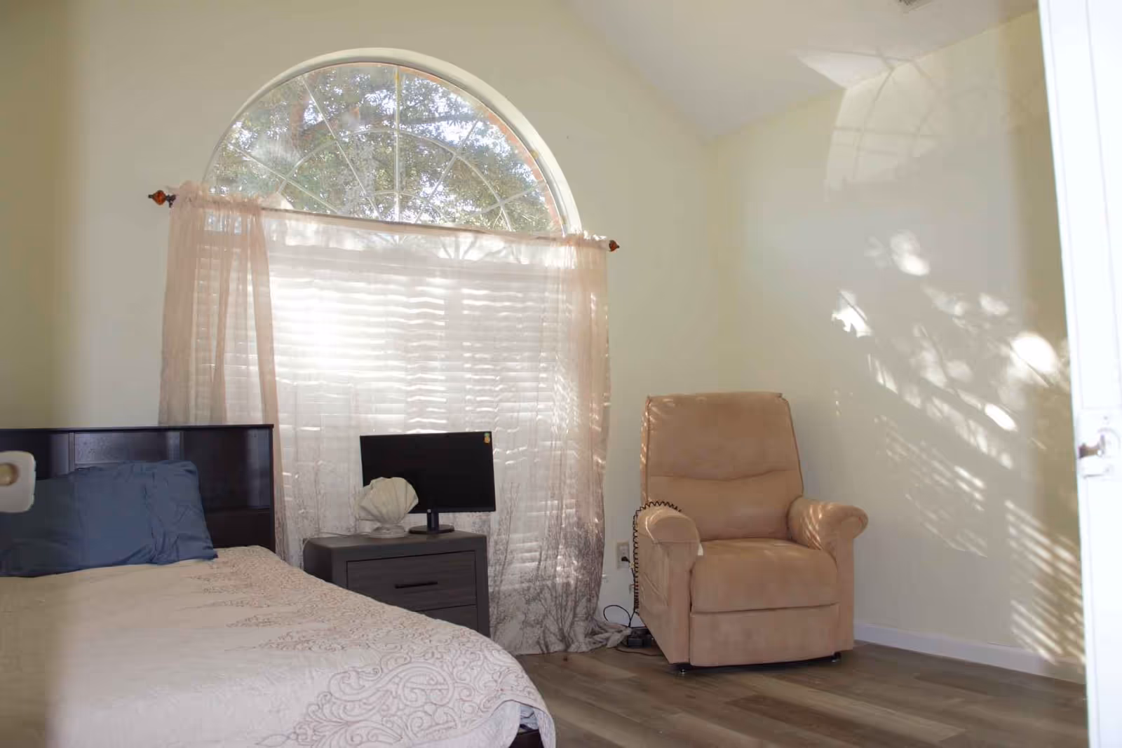 A bedroom with a bed on the left side covered with a beige patterned bedspread and a blue pillow. Next to the bed is a small gray nightstand with a small black monitor and a decorative shell. Behind the nightstand is a large arched window with sheer curtains allowing sunlight to filter through. On the right side of the room is a beige recliner chair. The floor is wooden and the walls are painted light cream.