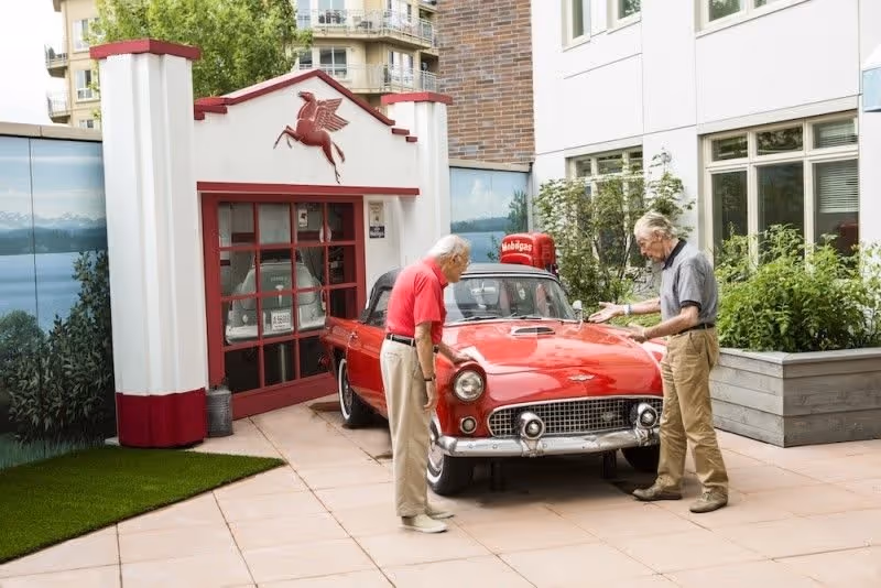 Two elderly men standing outside next to a vintage red car parked in front of a small white and red building with a Pegasus logo above the entrance. The setting appears to be a courtyard with greenery and windows of a building in the background.