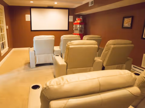 A cozy home theater room with several beige recliner chairs arranged in rows facing a large blank projection screen on a brown wall. There is a popcorn machine in the corner and framed artwork on the walls.
