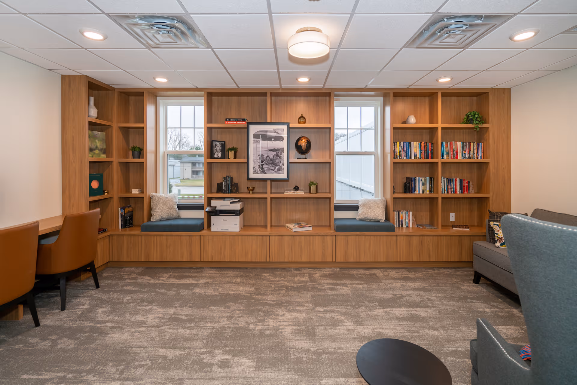 A bright communal sitting area with built-in wooden bookshelves, window seats, and armchairs.