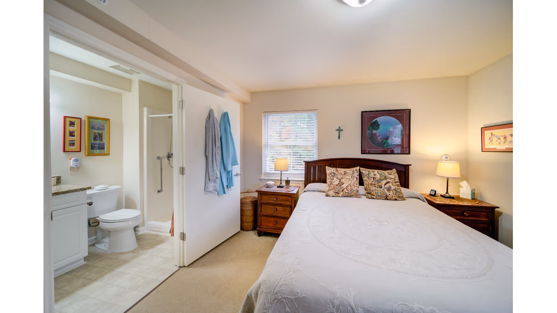 Well-lit bedroom with a bed, patterned pillows, two nightstands with lamps, and an open door showing an en-suite bathroom with a shower and toilet.