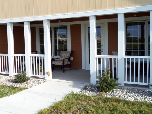 Outdoor porch area of a building with white railings and columns, two cushioned chairs, a concrete walkway, small bushes, and grass in the foreground.